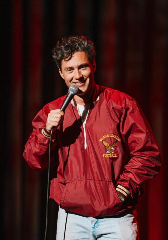 Alec wearing a red varsity style jacket holding a microphone on stage