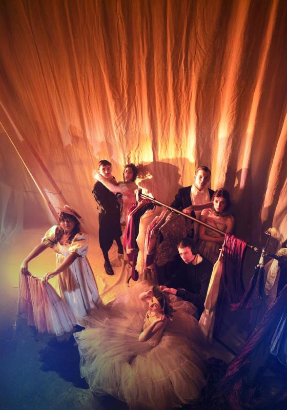 Backstage in a theatre,a group of performers in costumes look up to the camera in orange light
