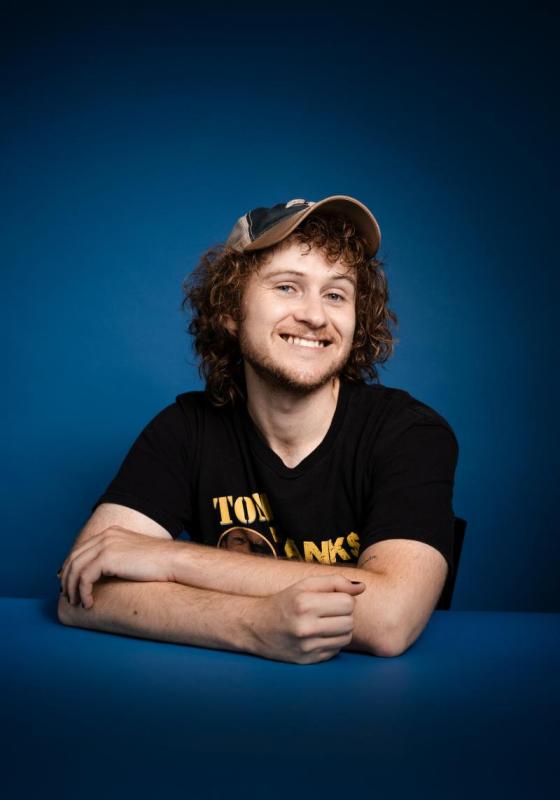 Casey smiles with arms loosely folded wearing a cap.