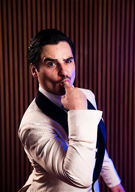 Joey wearing a white tuxedo sticks a finger in his mouth and raises an eyebrow