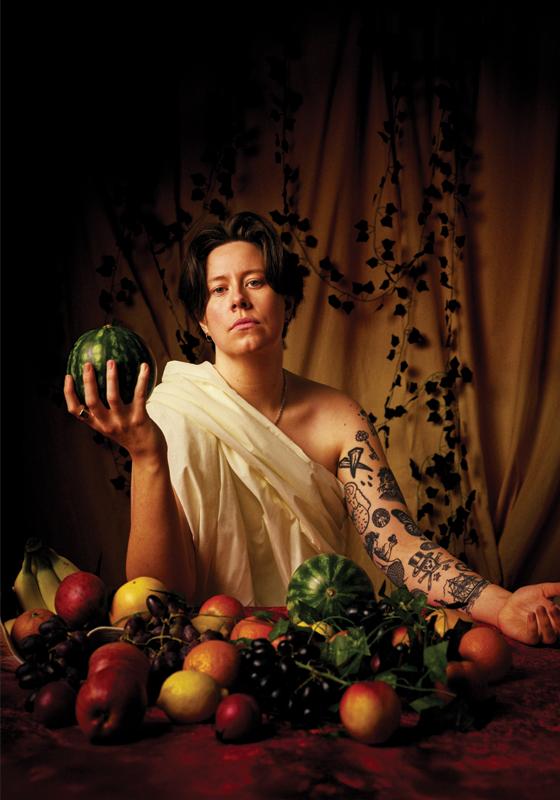 Chloe Petts wearing a toga amidst a lavish table of food and holding a watermelon in one hand