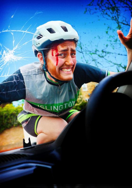 The performer dressed in a cycling outfit with a white helmet on top of a broken car windshield with blood running down their face