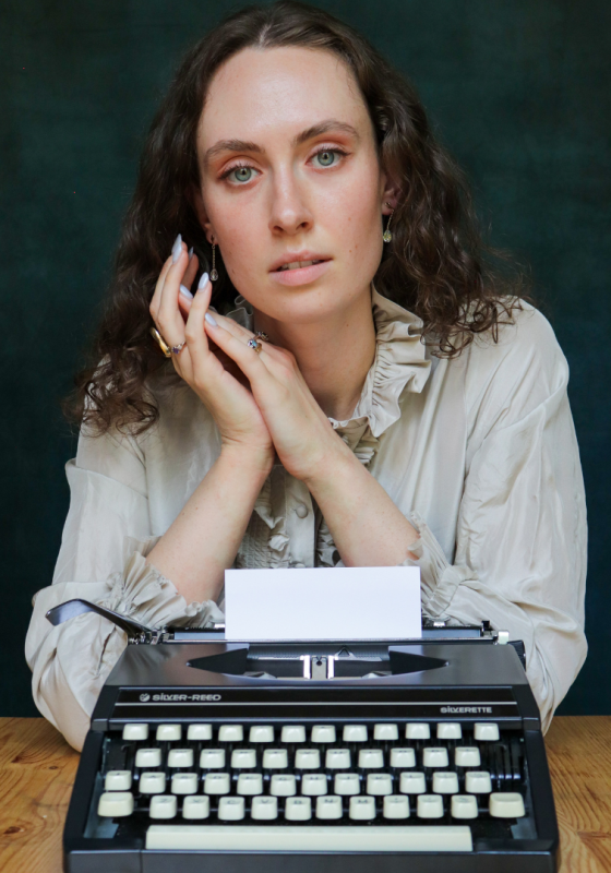 Woman sits behind a typewriter on a wooden desk, dark green background