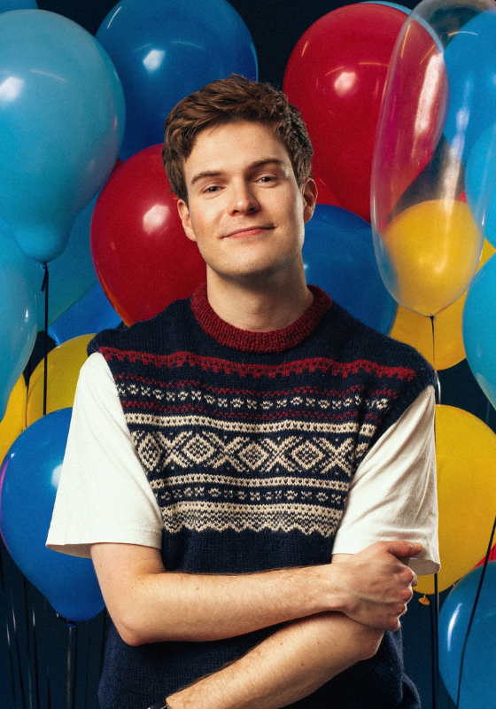 Thor stands in front of a big bunch of balloons wearing a knitted vest over a white t-shirt.