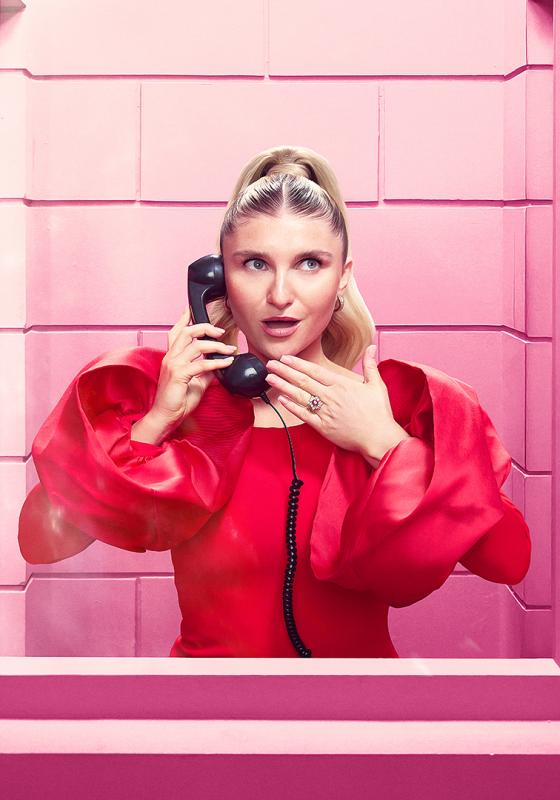 Sophie in a bright pink dress holds a phone standing behind glass.