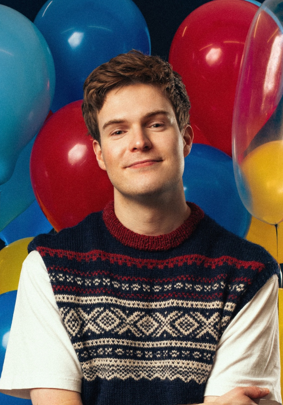 Thor stands in front of a big bunch of balloons wearing a knitted vest over a white t-shirt.