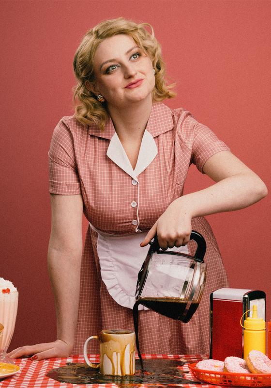 Lorna wearing a red gingham dress with an apron smiling sweetly as she pours coffee into an overflowing cup.