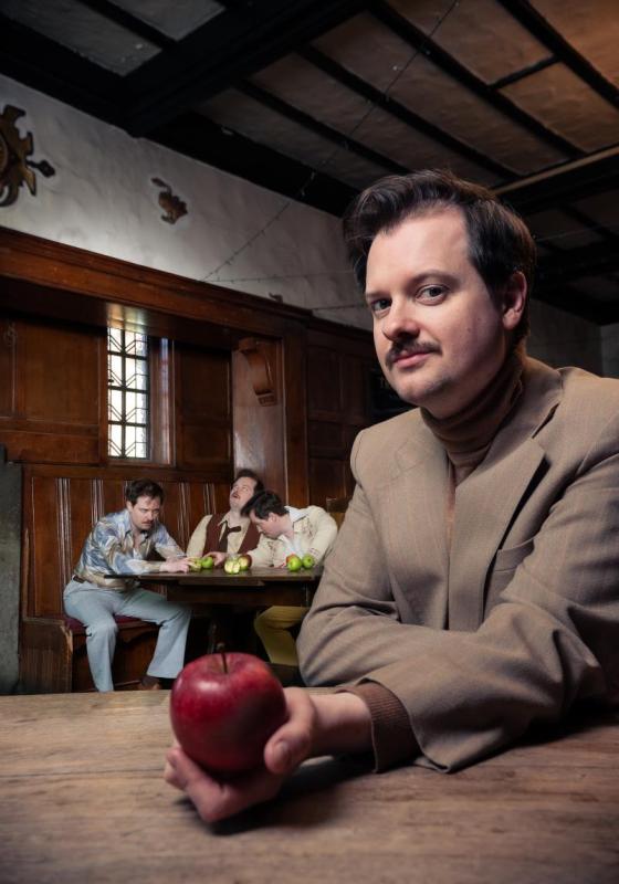 Andy in a pub wearing brown suit holding an apple at the forefront. In the background, Andy is cloned into three different drunk people.