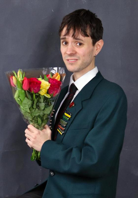 A boy in a school uniform blazer, and lots of achievement pins, holds a bunch of flowers and looks apologetic