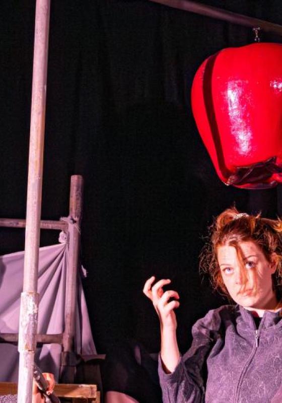 4 actors holding a metaphorical apple wearing grey jumpsuits whilst a large prop apple dangles above them.