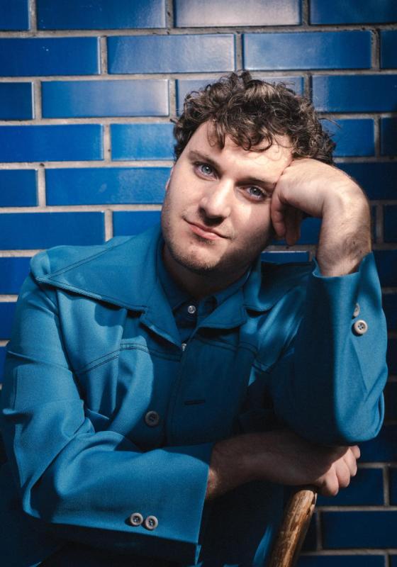 Headshot of performer wearing all blue set against a tiled blue background.