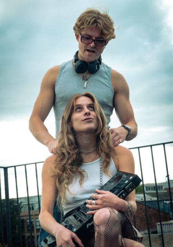A hetero couple sit on a balcony. The man looks down at the woman. He wears headphones round his neck, she holds DJ decks in hers. They look coy. 