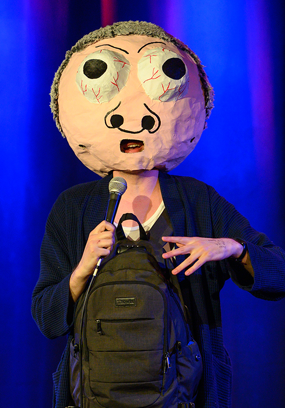 A papier mache head of Basil Crumbwick with bulging eyes and red veins worn by the performer holding a rucksack ad microphone.