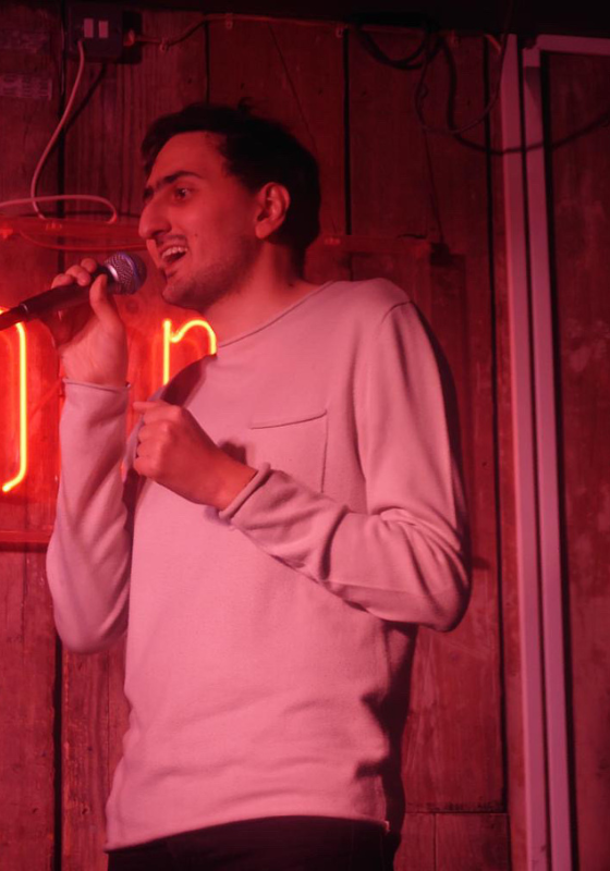Aarian holding a mic and doing stand-up in pink lighting