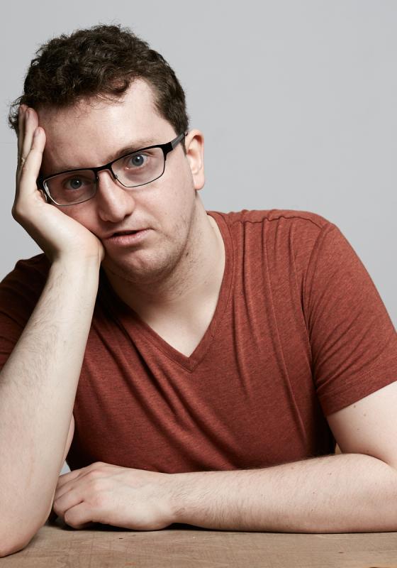 Jonny resting his face on his hands on a wooden table and a grey background.