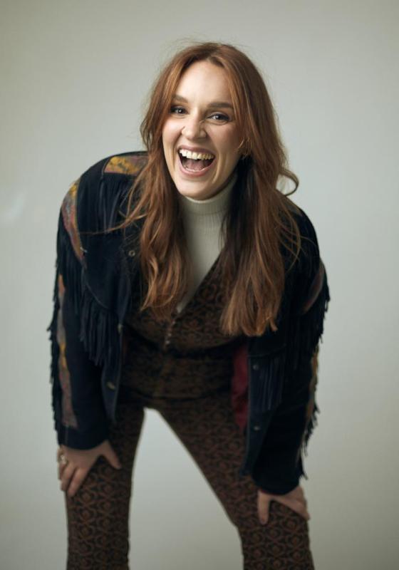 Katie Norris wearing a fringe jacket leans forward and laughs to camera