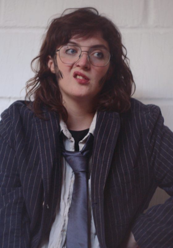 A photo Roger wearing a pinstripe suit and loose tie with sideburns and glasses.