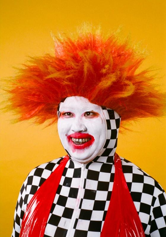 Su Mi with white face paint and red lipstick with a wild orange spiky wig. Wearing a full checkerboard one-piece with a red latex swimsuit.