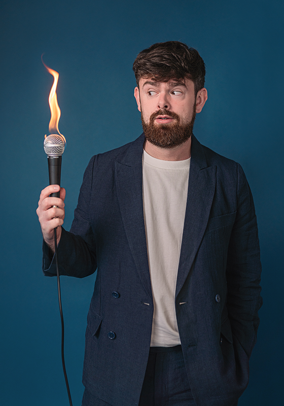 Tom, wearing a blue suit and white tshirt, holds a flaming mic in his right hand and is looking at it concerned. 