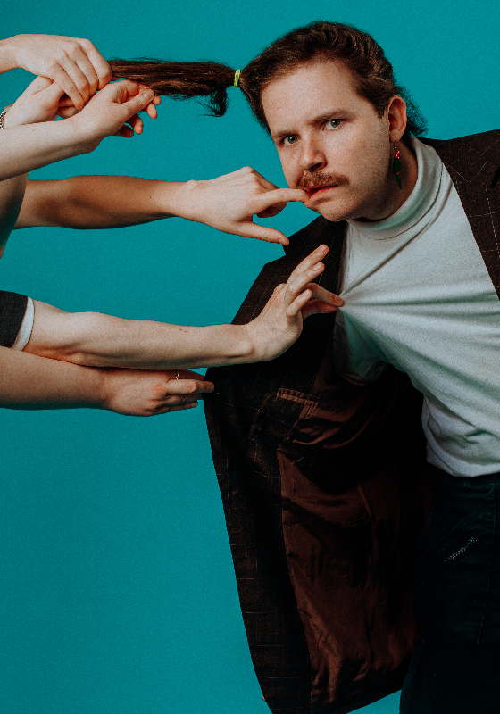 Performer in a suit is having their hair in a pony tail pulled by five different hands.