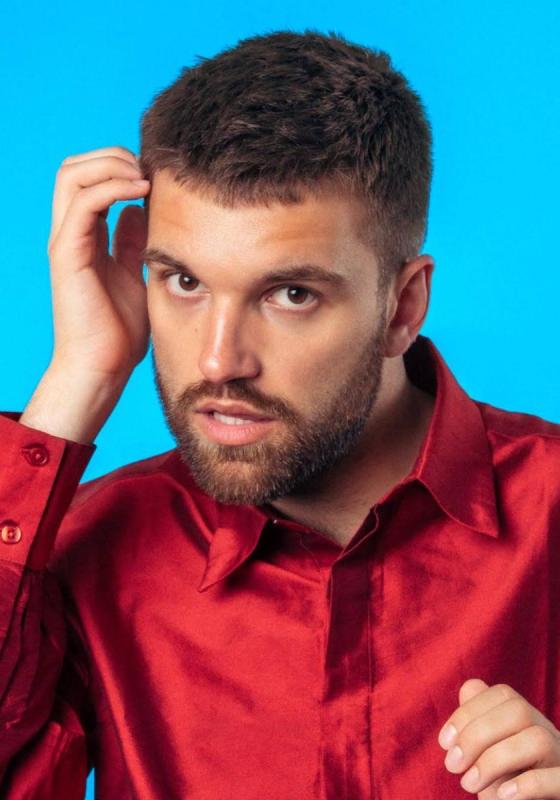Will in a bright red shirt against a vibrant blue backdrop lifting their hand to their head