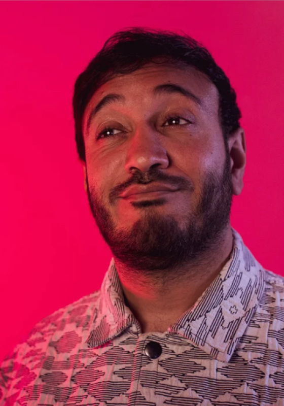 Bilal stands in front of a pink background, in a geometric shirt, looks with a wry smile into the distance