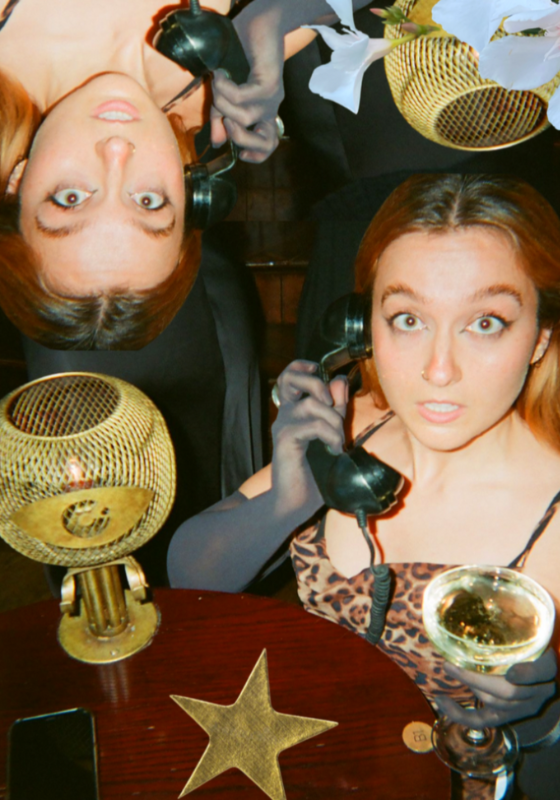 Em holding a black landline phone wearing black opera gloves and a leopard print dress. They hold a glass of champagne in their other hand.