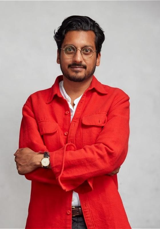 Photo of performer with arms loosely folded wearing an orange over shirt.