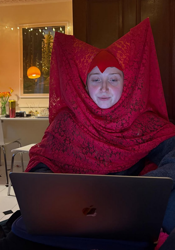 Bebe wearing a medieval inspired red headdress sat at a laptop.