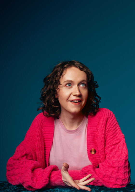 Dee sat at a table mid conversation wearing a pink shirt and cardigan..