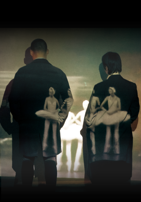 Two figures in suits stand with the faces to a wall. There are layers of photos on top including one of a ballet troupe.