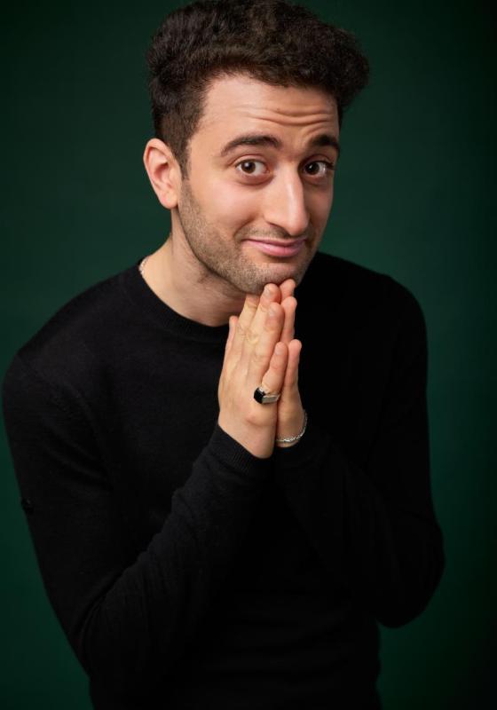 Hasan smiling at the camera with his hands together under his chin.