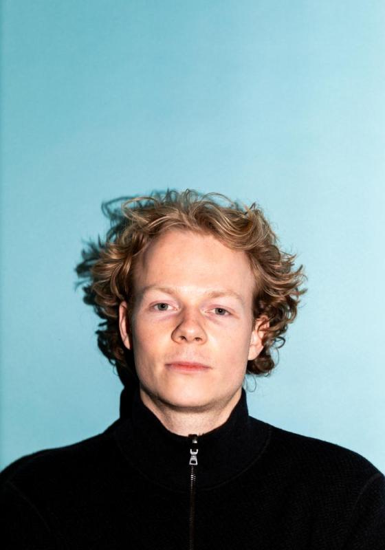 Alfie looks straight to camera, passport photo style, on a light blue backdrop wearing a black turtleneck.