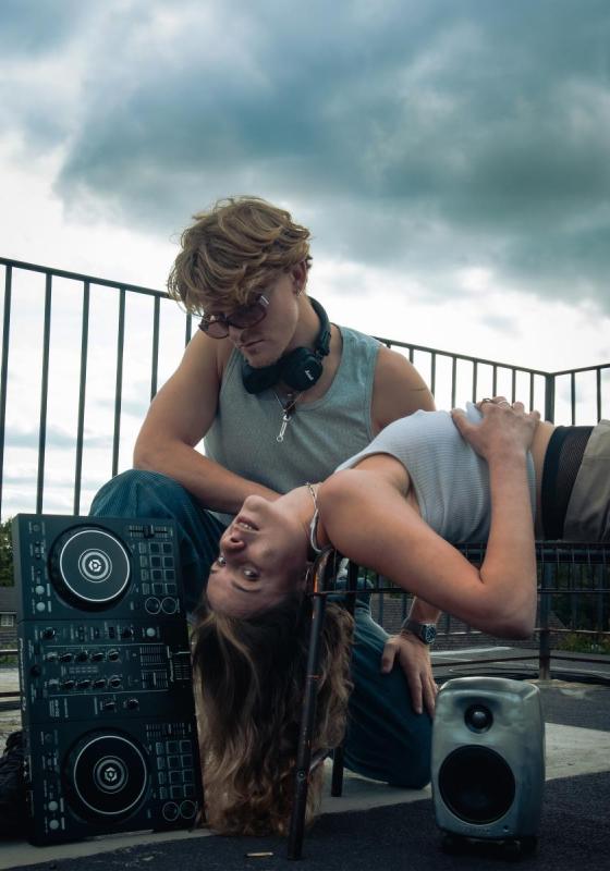 A woman lies down on a bench with a man behind her wearing headphones and holding a deck.