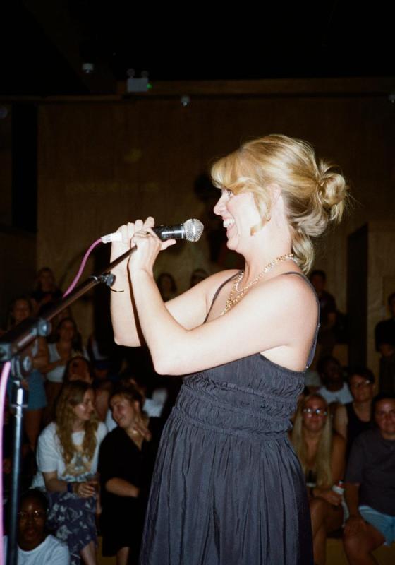 A woman with blonde hair stands on stage in front of a microphone, smiling with her hands clasped 