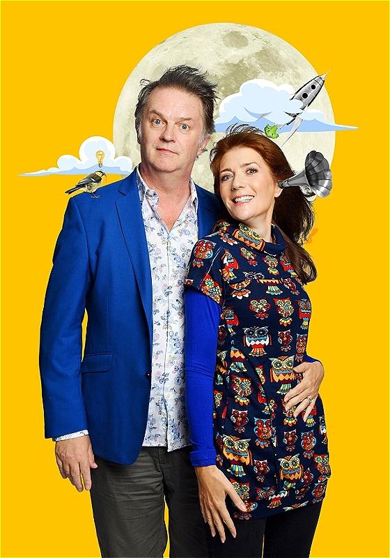 Paul and Suki stand looking at the camera in front of a tangerine backdrop and some clip art style photos of a big moon, rocket ship, cloud, small bird, etc