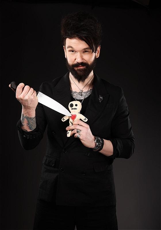 Bearded man dressed in black holds a dagger to the heart of a punky voodoo doll