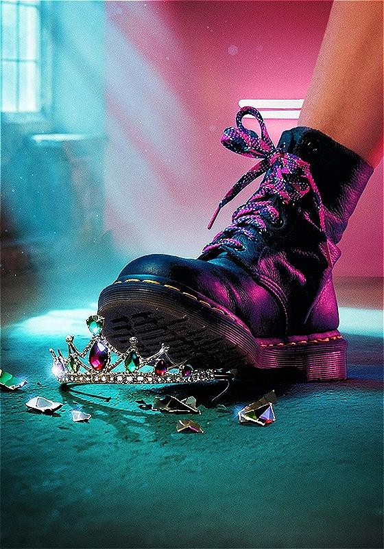 A black Dr. Martin boot stepping on and breaking a jewel-encrusted tiara under blue and pink lighting