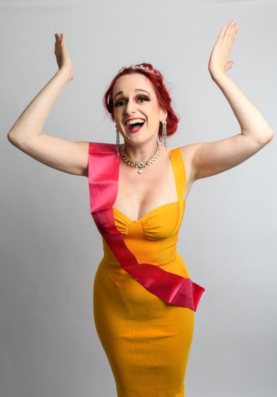 A person with red hair and elegant makeup, wearing a tiara, a bright yellow dress, pink sash, and jewelry, smiling joyfully with arms raised.