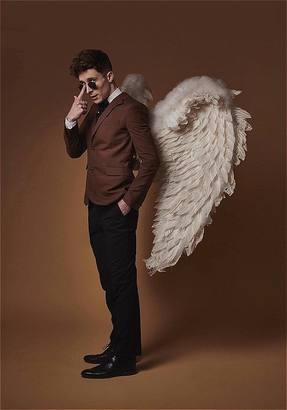 Larry is standing at a three-quarters turn to the camera, wearing angel wings, a brown business suit jacket, and round black sunglasses. He is pushing up the sunglasses with a finger. The background is brown.