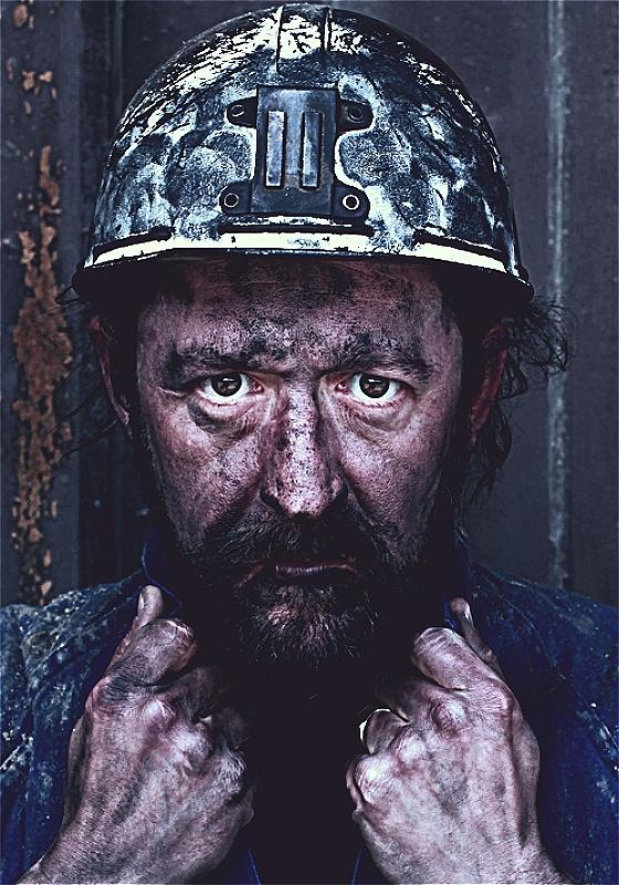 A man wearing a miner's outfit and covered in dust stares straight ahead before an iron background.