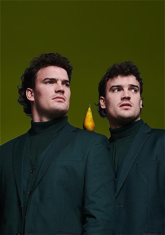 Two people in black stare moodily into the distance just off-camera. There is a pear on one person's shoulder in the middle of the photo.