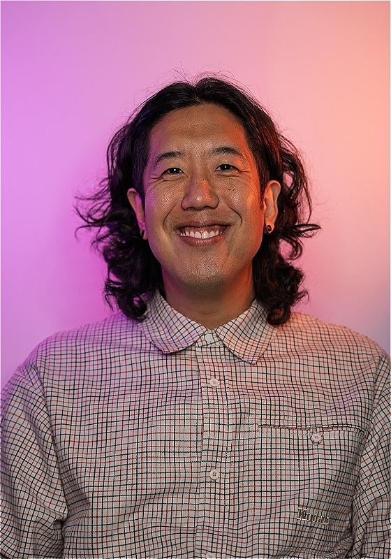 A smiling Harry wearing a checkered button-down shirt stands before a pastel purple and pink background.