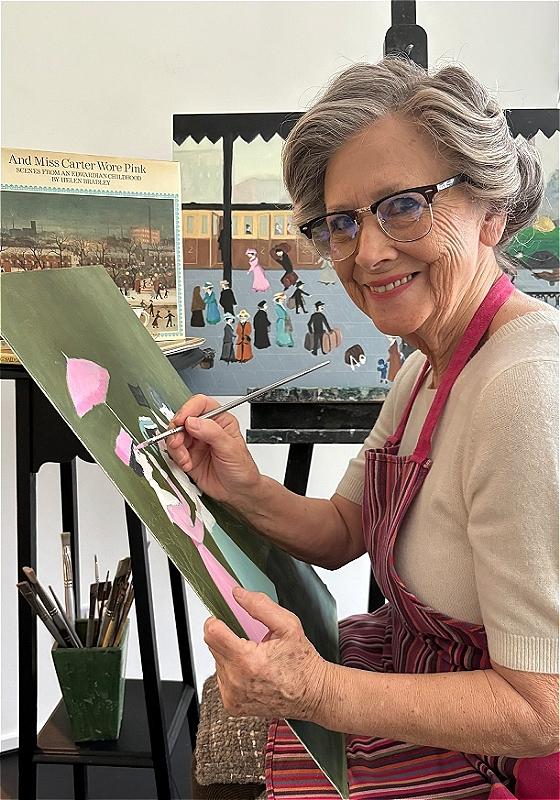 A person sits at an easel, painting, and smiles at the camera. Their grey hair is pinned up elegantly.