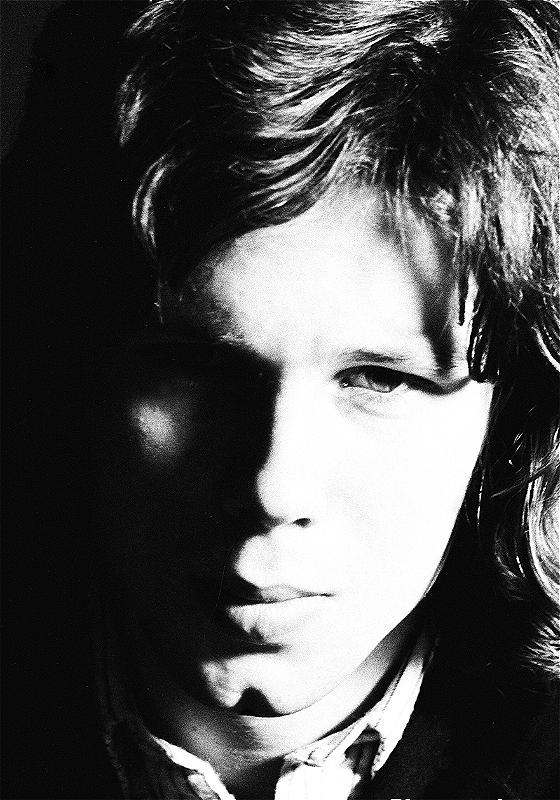 Black and white photo of a young man with shoulder-length hair staring straight ahead. 