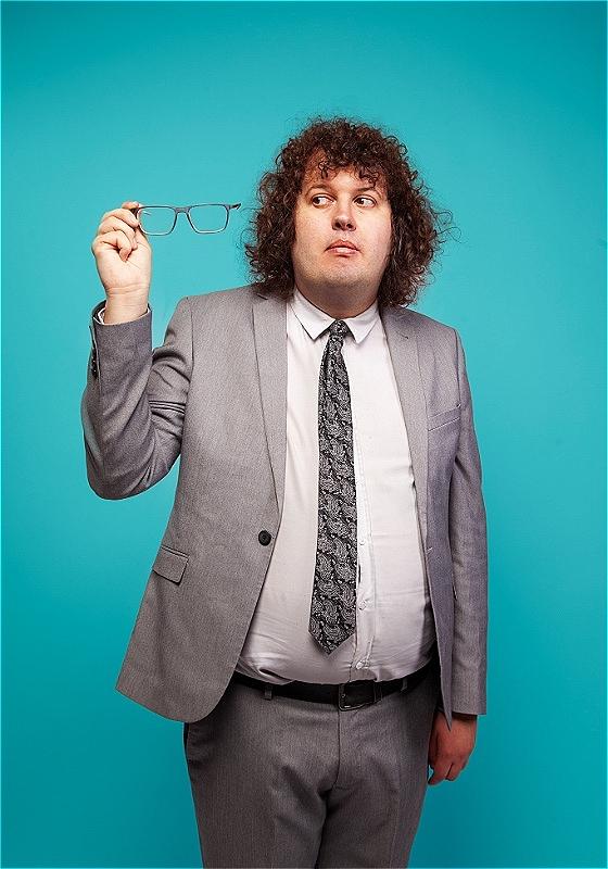 Ray is in a grey suit against a blue background, looking at their spectacles which they're holding up to the light in a gesture universally recognisable by people who use specs as 'is this still blurry'. They don't seem sure.