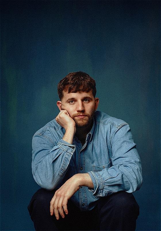 Rory is dressed all in blue against a blue background, crouching down and resting their chin on their hand and their elbow on their knee as they look at the camera. 