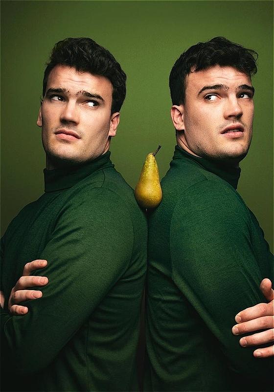 Two identical men wearing green turtleneck sweaters hold a pear between their shoulders before a green background.