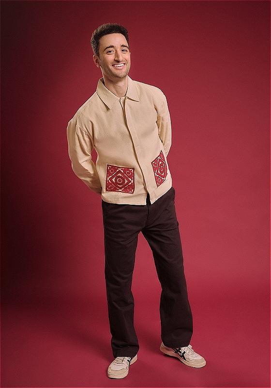 Hasan is smiling towards the camera with his hands crossed behind his back. His shirt has fabulous pockets embroidered in red patterns, matching the background.