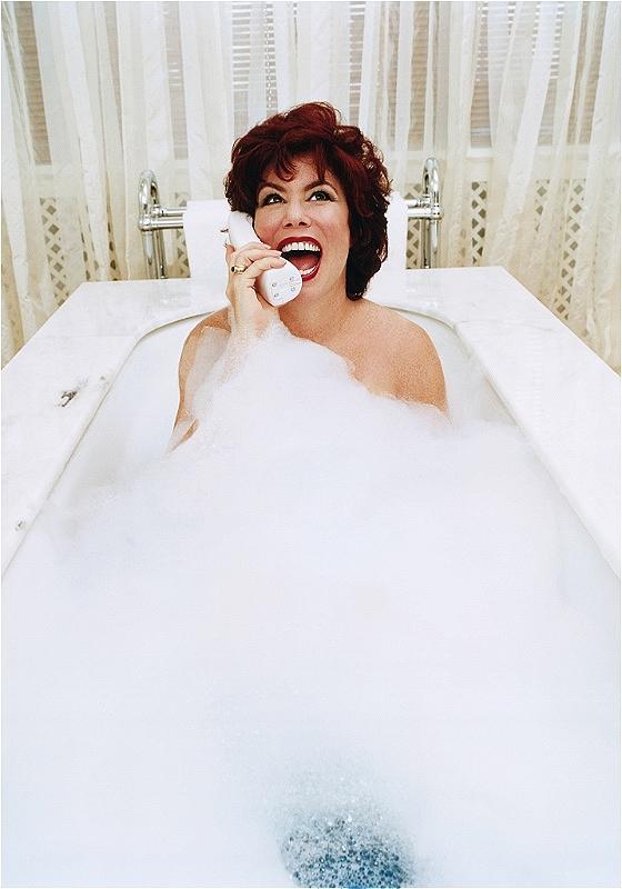 Ruby is in a luxurious, white-marble, bubble-filled bath. Bubbles cover her up to her shoulders, and she's holding an old-school white brick phone up to her ear and laughing. She's got fair skin, distinctive red hair, red lips, and a dazzling smile. 